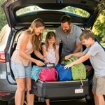 Équipements pour voyager en voiture avec des enfants : 12 essentiels Une famille souriante range des valises et des sacs organisés dans le coffre ouvert d’une voiture avant de partir en vacances.