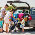 Une famille charge des valises, une poussette pliée et des sacs de plage dans le coffre ouvert d’un break d’occasion, sur un parking lumineux avant les vacances.