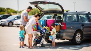 Une famille charge des valises, une poussette pliée et des sacs de plage dans le coffre ouvert d’un break d’occasion, sur un parking lumineux avant les vacances.